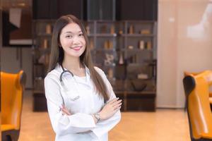 médico mulher asiática sorriso permanente de bom humor com os braços cruzados na sala de escritório no hospital. vestindo uma túnica branca e estetoscópio. foto