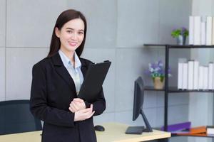 jovem profissional de negócios asiáticos de terno preto sorri alegremente enquanto ela trabalha e mantém a área de transferência com confiança no escritório. foto