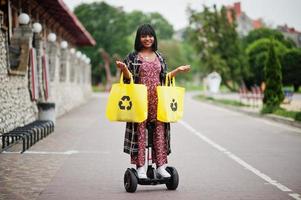 linda mulher afro-americana usando segway ou hoverboard. garota negra com símbolo de reciclagem de sacos ecológicos de pano amarelo. foto