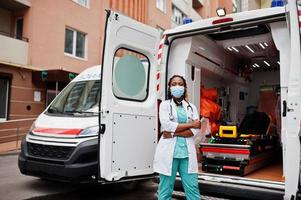 paramédica afro-americana em máscara médica protetora facial em frente ao carro da ambulância. foto