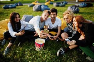 jovem grupo multiétnico de pessoas assistindo filme no poof no cinema ao ar livre e fazendo selfie no telefone. foto