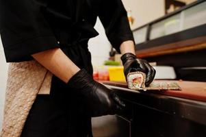 Feche as mãos do chef profissional vestindo luvas pretas fazendo sushi e pãezinhos em uma cozinha de restaurante de comida tradicional japonesa. foto