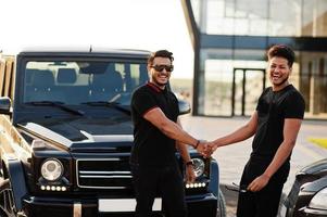 dois irmãos asiáticos usam todo preto posando perto de carros suv e apertando as mãos. foto