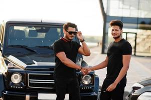 dois irmãos asiáticos usam todo preto posando perto de carros suv e apertando as mãos. foto