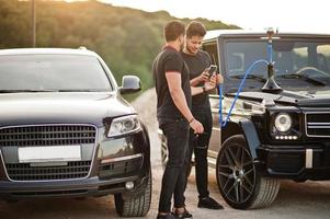 dois irmãos asiáticos usam todo preto posando perto de carros suv e fumam cachimbo de água. foto