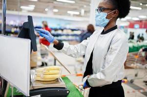 mulher africana usando máscara médica descartável e luvas fazendo compras no supermercado durante o surto de pandemia de coronavírus. fêmea negra pesa frutas em época epidêmica. foto