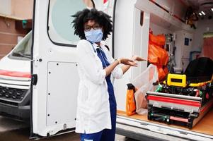 paramédica afro-americana em máscara médica protetora facial em frente ao carro da ambulância. foto