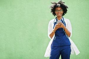 retrato de médica afro-americana com estetoscópio vestindo jaleco, falando por telefone celular. foto