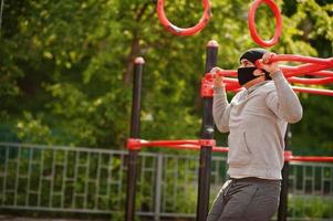 retrato esportivo homem árabe com máscara facial médica preta fazendo exercícios de treino no local da academia ao ar livre durante a quarentena de coronavírus. foto