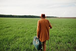 homem elegante de óculos, jaqueta marrom e chapéu com bolsa posou em campo verde. foto