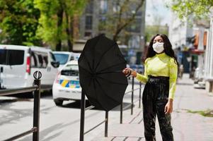 mulher afro-americana posando com máscara facial para proteger de infecções de bactérias, vírus e epidemias e segurar o guarda-chuva contra o carro da polícia. foto