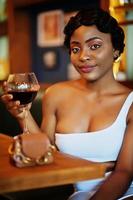 mulher afro-americana, penteado retrô vestido branco no restaurante com copo de vinho. foto