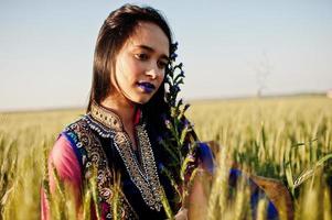 tenra garota indiana em saree, com lábios violeta compõem posou no campo no pôr do sol. modelo elegante da índia. foto