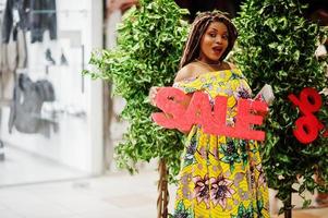 linda garota afro-americana de pequena altura com dreadlocks, use um vestido amarelo colorido, segurando a carta de sinal de venda no shopping center. foto