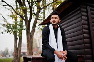 homem elegante indiano em roupas tradicionais pretas com lenço branco posou ao ar livre contra um fundo de madeira. foto