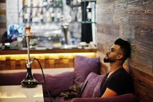 homem árabe de barba elegante de óculos e camiseta preta fumando narguilé bar interno. modelo árabe descansando. foto