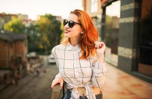 mulher ruiva atraente em óculos de sol, use blusa branca posando na rua contra o edifício moderno. foto