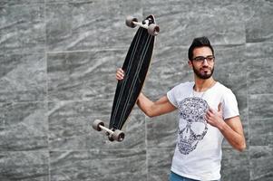 homem árabe de estilo de rua em óculos com longboard posou contra a parede cinza. polegar para cima mostra. foto