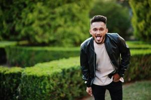 homem de barba indiana elegante na jaqueta de couro preta mostra tonque. modelo da índia posou no jardim verde da cidade. foto