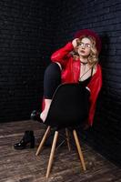 retrato de estúdio de menina loira de chapéu vermelho, óculos e jaqueta de couro posou na cadeira contra a parede de tijolos. foto
