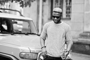 menino afro-americano elegante na camisola cinza e óculos de sol pretos posou na rua contra o carro amarelo. homem de negócios preto na moda. foto