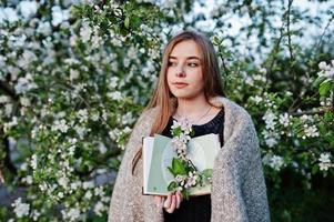 jovem morena na manta contra árvore de flor de primavera e segurando o livro nas mãos. foto