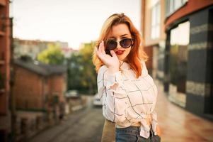 mulher ruiva atraente em óculos de sol, use blusa branca posando na rua contra o edifício moderno. foto