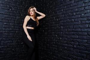 menina morena bonita usa preto, posando no estúdio contra a parede de tijolos escuros. retrato de modelo de estúdio. foto