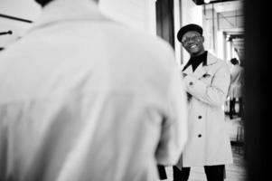 homem afro-americano casual elegante na boina preta e capa de chuva branca na loja de roupas de provador, olhando para o espelho. foto