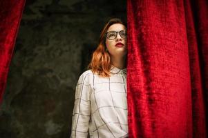 mulher ruiva atraente em óculos, use blusa branca posando no arco de cortinas vermelhas abertas. foto