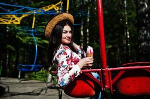 retrato de menina morena de óculos cor de rosa e chapéu com sorvete no parque de diversões. foto