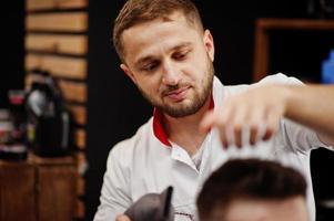 jovem barbudo cortando o cabelo pelo cabeleireiro enquanto está sentado na cadeira na barbearia. alma de barbeiro. foto