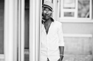 homem afro-americano elegante de camisa branca e calça colorida com chapéu e óculos posou ao ar livre na cabine telefônica. menino modelo preto na moda. foto