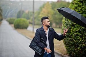 homem de barba árabe alto na moda usa casaco preto com guarda-chuva e bolsa posada no dia de chuva. foto