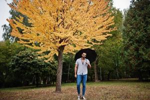 homem de barba árabe alto na moda usar camisa, jeans e óculos de sol posou no parque outono com guarda-chuva contra árvore de folhas amarelas. foto