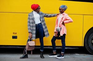 duas jovens mulheres muçulmanas africanas elegantes, atraentes, altas e magras em hijab ou lenço de cabeça de turbante e casaco posaram contra ônibus amarelo. foto