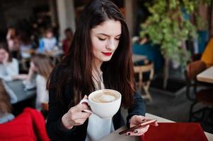 menina morena sentada no café com uma xícara de cappuccino, trabalhando com laptop vermelho e olhando para o celular. foto