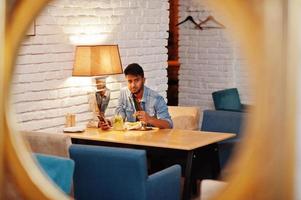 retrato de bonito bem sucedido barbudo sul-asiático, jovem indiano freelancer em camisa jeans azul sentado no café com nuggets de frango e limonada. foto