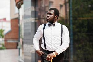 homem afro-americano elegante bonito com roupa formal, gravata borboleta e suspensórios, bengala. foto