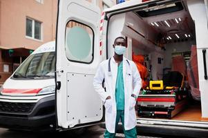 paramédico masculino africano em máscara médica protetora facial em frente ao carro da ambulância. foto