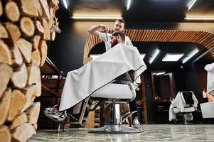 jovem barbudo cortando o cabelo pelo cabeleireiro enquanto está sentado na cadeira na barbearia. alma de barbeiro. foto