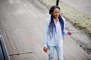 menina afro-americana elegante com dreads segurando o celular na mão, clima de neve ao ar livre. foto