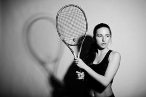 retrato preto e branco do jogador de mulher jovem e bonita em roupas esportivas, segurando a raquete de tênis em pé contra um fundo branco. foto