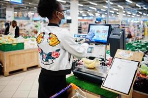 mulher africana usando máscara médica descartável e luvas fazendo compras no supermercado durante o surto de pandemia de coronavírus. mulher negra escolhe bananas em época epidêmica. foto