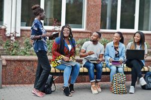 grupo de cinco estudantes universitários africanos passando tempo juntos no campus no pátio da universidade. amigos afro negros estudando no banco com itens escolares, notebooks laptops. foto