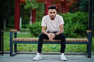 homem de barba indiana elegante na camiseta rosa. modelo da índia sentado no banco da cidade. foto