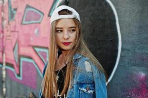 menina elegante casual hipster em boné e jeans usa música de fones de ouvido do telefone celular contra grande parede de graffiti com bomba. foto