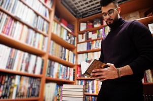 homem alto estudante árabe inteligente, use gola alta violeta e óculos, na biblioteca com pilha de livros.