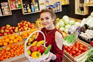 garota de vermelho segurando diferentes frutas e legumes na cesta na loja de frutas. foto