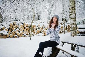 linda garota encaracolada de suéter e chapéus sentado no banco no parque florestal nevado no inverno. foto